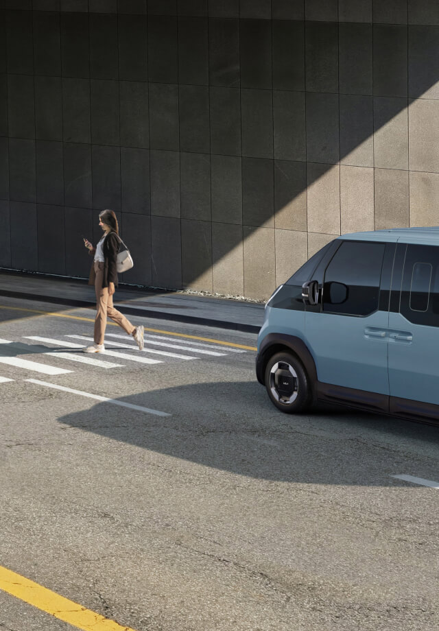 Der Kia PV5 Passenger hält am Zebrastreifen, während ein Fußgänger vorbeigeht.